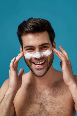 Temiz ciltli ve beyaz maskeli neşeli bir adam mavi arka planda izole edilmiş kameraya gülümseyen gözenekleri temizlemek için burnuna sürmüş.
