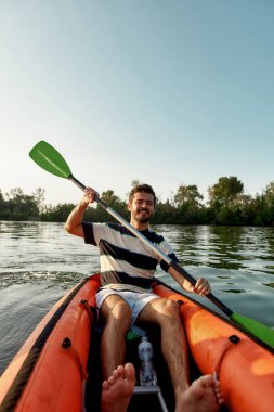 Aktif genç bir adam, bir yaz öğleden sonrasında, etrafında huzurlu bir doğa olan gölde kano sürerken neşeli görünüyor.