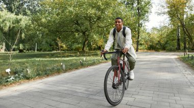 Sırt çantalı bir adam park yolu boyunca bisiklet sürüyor.