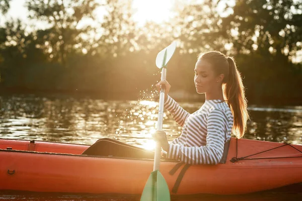 Vista lateral de la joven caucásica remando kayak en un lago rodeado de hermosa naturaleza al atardecer — Foto de Stock