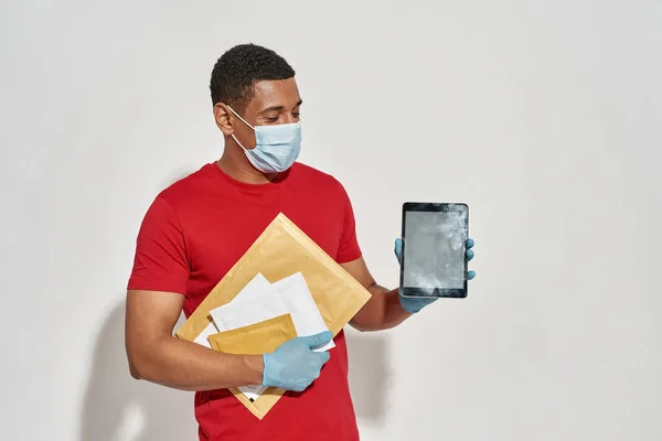 Donáška muž v ochranných rukavicích drží balíčky a písmena, ukazuje tablet PC na kameru, stojící izolované přes světle šedé pozadí — Stock fotografie