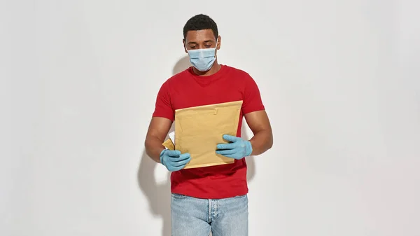 Donáška muž s obličejovou maskou a ochranné rukavice držení, při pohledu na balíčky a obálky při pózování izolované přes světle šedé pozadí — Stock fotografie