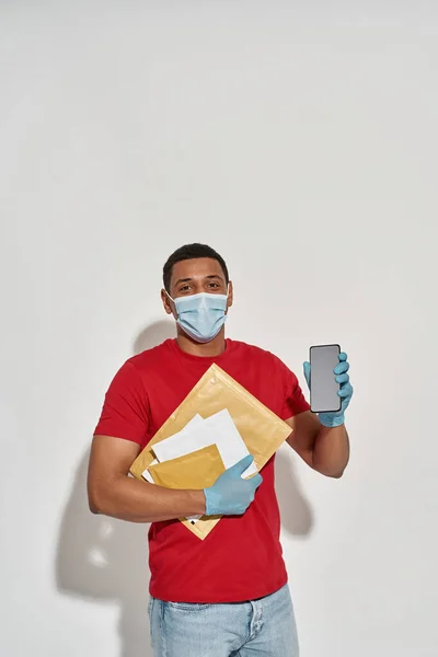 Repartidor con mascarilla facial y guantes protectores mirando a la cámara, sosteniendo paquetes, cartas y banca móvil de teléfonos inteligentes, posando aislado sobre fondo gris claro —  Fotos de Stock