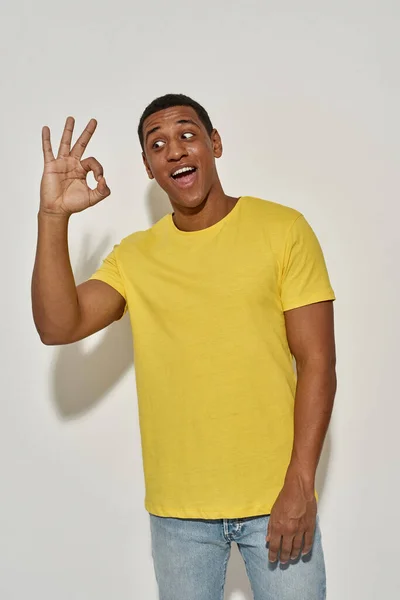 Jovem brincalhão em camiseta amarela casual olhando emocional e mostrando gesto sinal OK ao posar, de pé isolado sobre fundo cinza — Fotografia de Stock