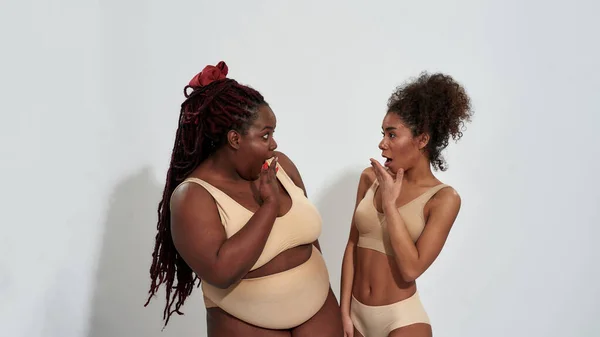 Plusz méret és vékony afro-amerikai nők fehérneműben néztek egymásra meglepődve, együtt álltak elszigetelve szürke háttér felett — Stock Fotó