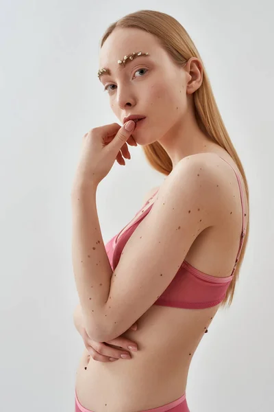 Креативний знімок красивої молодої кавказької жіночої моделі з квітами в брови в рожевому прозорому бюстгальтері торкається її губ, дивлячись на камеру, позує ізольовано на світло-сірому фоні — стокове фото