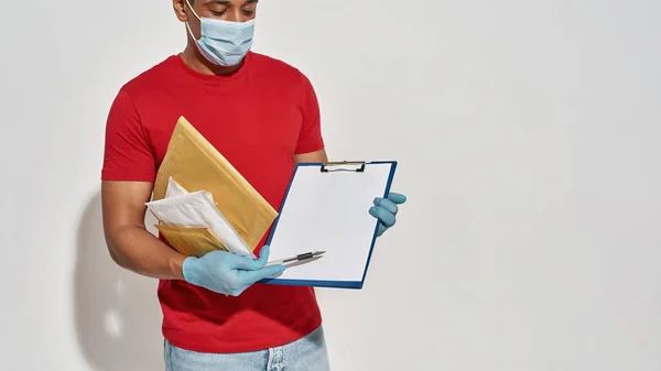 Cropped shot of delivery man wearing face mask and protective gloves holding parcels and envelopes, showing form for signing on clipboard, standing isolated over light gray background — Stock Photo, Image