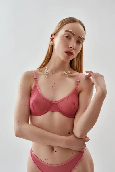 Delgado joven modelo femenino caucásico con flores en las cejas y en las clavículas con ropa interior rosa transparente de pie, posando aislado sobre fondo gris claro —  Fotos de Stock