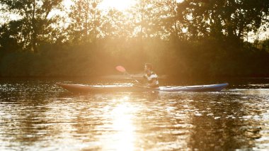 Genç adam elinde bir kürek tutuyor, gün batımında doğa ile çevrili bir gölde kayak yapıyor.