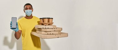 Koruyucu maske takan erkek kurye ve eldivenler kameraya bakıyor, akıllı telefon bankacılığı, pizza kutuları ve içkiler gri arka planda izole edilmiş.