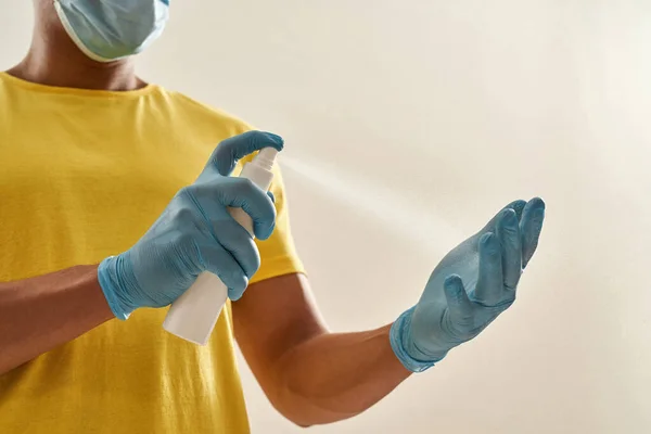 Beskuren skott av leverans man i gul t-shirt uniform steril ansiktsmask och handskar sprutning sanitetsrengöringsmedel, desinficera handskar isolerade över grå bakgrund — Stockfoto