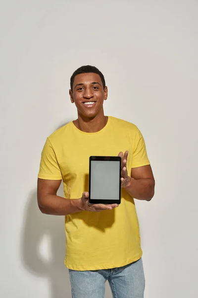 Joyeux jeune homme en t-shirt jaune décontracté souriant à la caméra, montrant ordinateur tablette numérique avec écran blanc tout en se tenant isolé sur fond gris — Photo