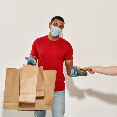 Koruyucu maskeli ve eldivenli mutlu teslimatçı. Bir sürü kahverengi el işi kâğıt torbasıyla poz veriyor, ödeme terminalini tutuyor. Müşteri ise açık gri arka planda kredi kartıyla ödeme yapıyor.