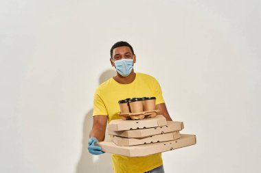 Koruyucu yüz maskesi takan neşeli erkek kurye kameraya bakıyor, siparişleri tutuyor, gri arka planda tek başına duruyor.