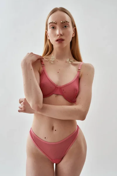 Sensual modelo feminino caucasiano jovem com flores em suas sobrancelhas e nas clavículas vestindo rosa transparente roupa interior em pé, posando isolado sobre fundo cinza claro — Fotografia de Stock