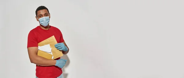 Young delivery man wearing face mask and protective gloves looking at camera, holding parcels and envelops, posing isolated over light gray background — Stock Photo, Image