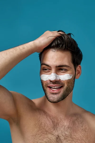 Sexy shirtloze man met wit masker aangebracht op de neus voor het reinigen van poriën glimlachen opzij, aanraken van zijn bruine haar geïsoleerd over blauwe achtergrond — Stockfoto