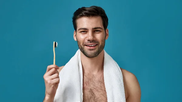 Close-up portret van vrolijke kerel met handdoek om zijn nek glimlachen op camera, vasthouden tandenborstel terwijl poseren geïsoleerd over blauwe achtergrond — Stockfoto
