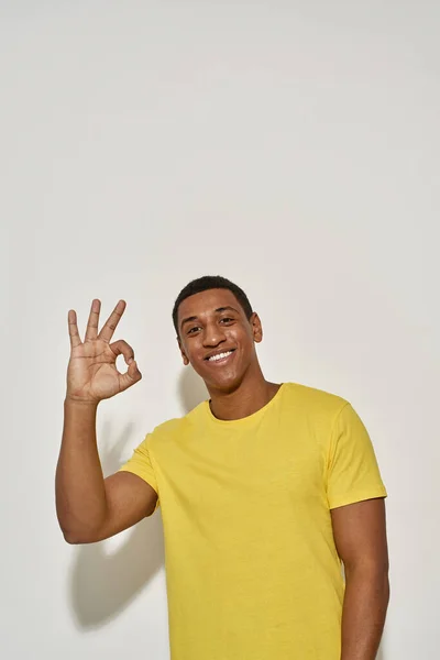 Happy young man in casual yellow t shirt looking at camera and showing OK sign gesture while posing, standing isolated over gray background — Stok fotoğraf