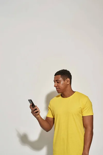 Focalizzato giovane uomo in casual t shirt gialla cercando occupato, tenendo e utilizzando il suo smartphone mentre in piedi isolato su sfondo grigio — Foto Stock