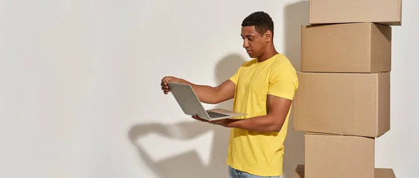 Professional male mover in yellow t shirt using laptop, standing near a stack of cardboard boxes while doing home relocation — Fotografia de Stock