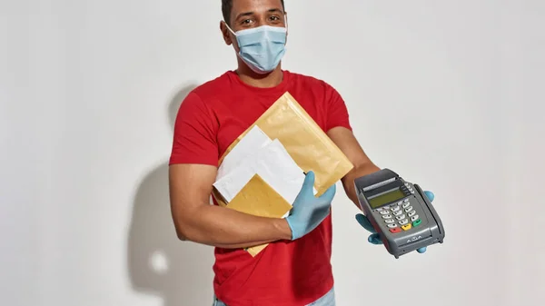 Cropped shot of delivery man in face mask and protective gloves holding parcels, envelopes and payment terminal for paying with credit card isolated over light gray background — Stock Fotó