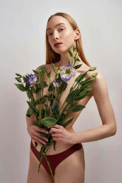 Креативний знімок натуральної молодої кавказької половини гола жінка дивиться на камеру, тримаючи квіти еустоми над її грудьми, позує ізольовано на сірому фоні — стокове фото