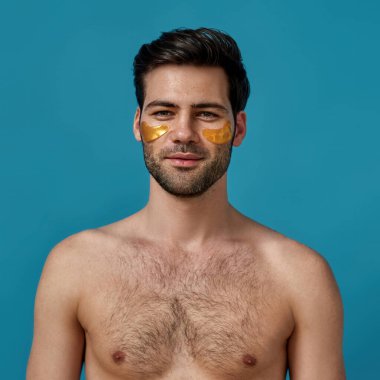 Shirtless brunette guy with golden gel under eye patches applied on his face smiling at camera, posing isolated over blue background