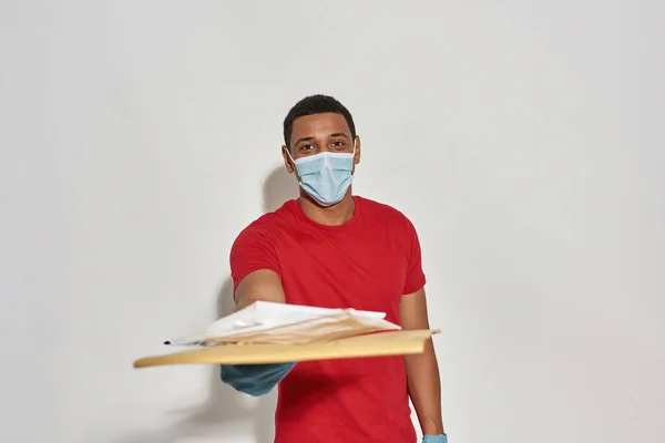 Cheerful delivery man wearing face mask and protective gloves smiling at camera, reaching out a hand while giving parcels and envelops, standing isolated over light gray background — Stock Fotó