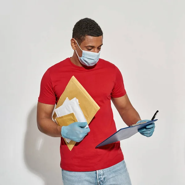Busy delivery man wearing face mask and protective gloves looking at clipboard, holding parcels and envelopes while posing isolated over light gray background — Stock Photo, Image