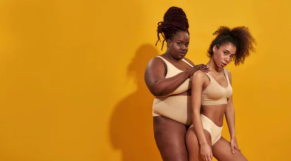 Two confident african american women in beige underwear with different body size posing together isolated over yellow background —  Fotos de Stock