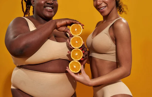 Cropped shot of two african american women in underwear smiling while holding halves of ripe juicy orange, standing isolated over orange background — Zdjęcie stockowe