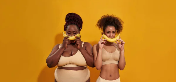 Dvě veselé mladé africké Američanky ve spodním prádle drží zralé žluté banány nad ústy jako úsměv, pózují izolované nad oranžovým pozadím — Stock fotografie