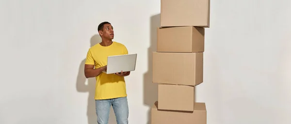 Hombre ocupado moviendo en camiseta amarilla mirando a un lado, sosteniendo el ordenador portátil, de pie cerca de una pila de cajas de cartón mientras que hace la reubicación del hogar —  Fotos de Stock