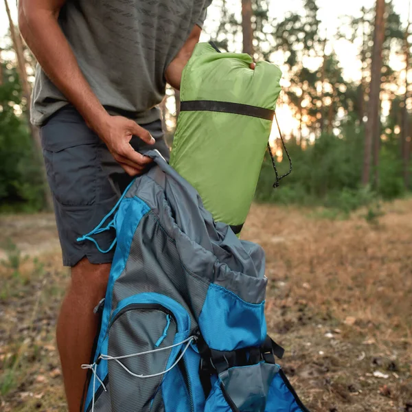 Cropped shot of young male tourist unpacking sleeping bag and other camping equipment, standing in the summer forest — Stock Photo, Image