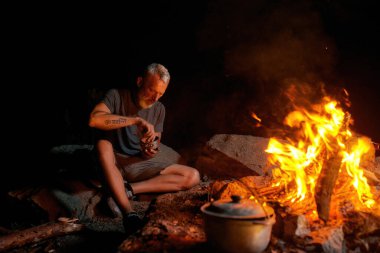 Olgun bir adam, vahşi doğada kamp ateşinin yanında tek başına oturmuş yemek yiyen bir turist.