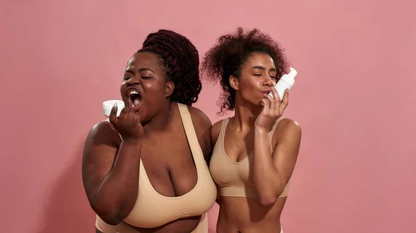 Imagen de una mujer afro de talla grande tratando de comer una crema y su amiga está besando una botella — Foto de Stock
