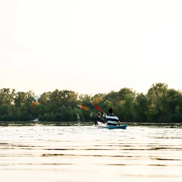 Rear view of two young guys best friends paddling kayak on a lake surrounded by peaceful nature