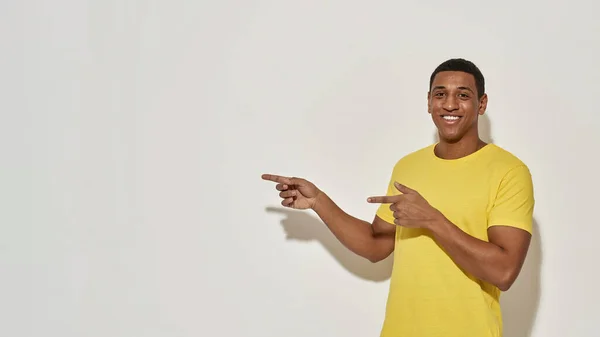 Jovem alegre em camiseta amarela casual sorrindo para a câmera e apontando os dedos indicadores para o lado enquanto posando isolado sobre fundo cinza — Fotografia de Stock