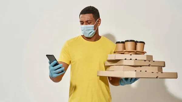 Mensajero masculino con máscara protectora y guantes usando la banca móvil de teléfonos inteligentes mientras lleva cajas de pizza y bebidas aisladas sobre fondo gris —  Fotos de Stock