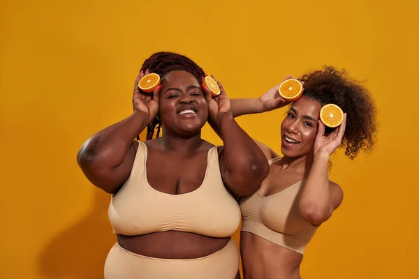 Due giovani donne africane americane eccitate che tengono metà di arancia matura succosa, posando insieme isolate su uno sfondo arancione — Foto Stock