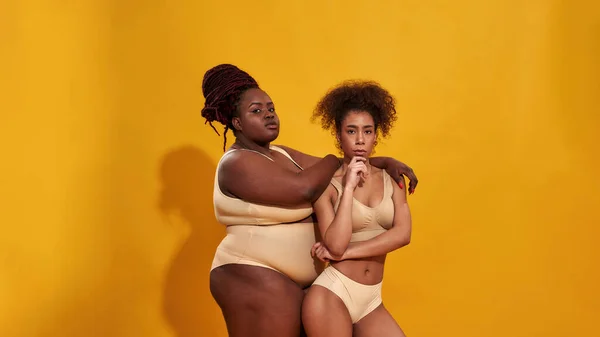 Two serious african american women in beige underwear with different body size standing together, posing isolated over yellow background — Stock Photo, Image