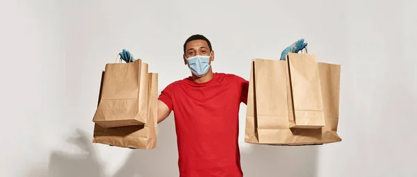 Cheerful delivery man in protective mask and gloves looking at camera, showing many brown craft paper bags for takeaway, posing isolated over light gray background — Stock Photo, Image