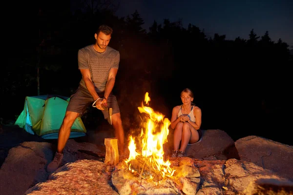 Un par de jóvenes acampan en el bosque. Hombre cortando leña para un fuego con un hacha — Foto de Stock