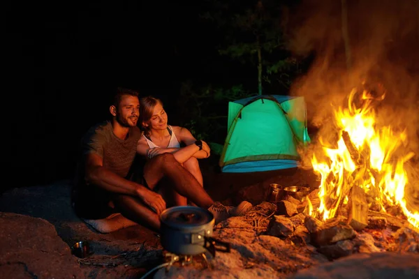 Pareja joven de turistas buscando relajados mientras acampan, sentados cerca de la fogata, cocinando la cena en la naturaleza — Foto de Stock