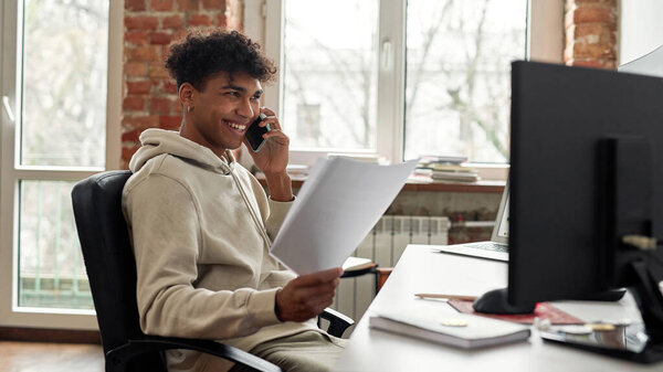 Успешный молодой мужчина-трейдер с бумагами, улыбающийся во время разговора по телефону с клиентом, сидящий за столом, торгующий из дома