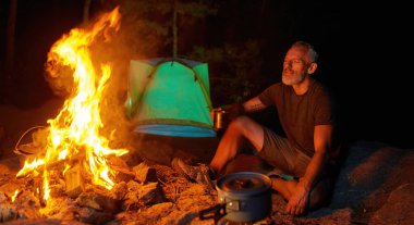 Rahat erkek turist sıcak çay ya da kahve içerken geceleri ormanda kamp ateşinin yanında oturuyor.