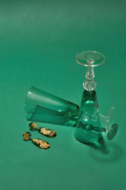 İçinde altın folyo bulunan epoksi reçineden yapılmış el yapımı sarkık küpeler ve yeşil arka planda izole edilmiş iki şarap kadehi.