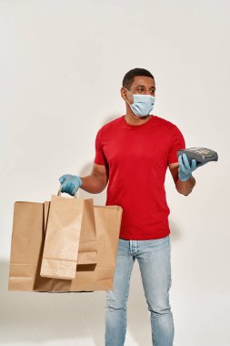 Kredi kartıyla ödeme yapan koruyucu maskeli ve eldivenli teslimatçı, açık gri arka planda izole edilmiş paket servis için bir sürü kahverengi el çantası ile ayakta duruyor.