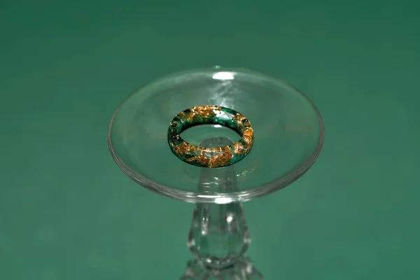 Tiro criativo de anel artesanal elegante feito de resina epóxi com folha de ouro no interior em vidro de vinho isolado sobre fundo verde — Fotografia de Stock